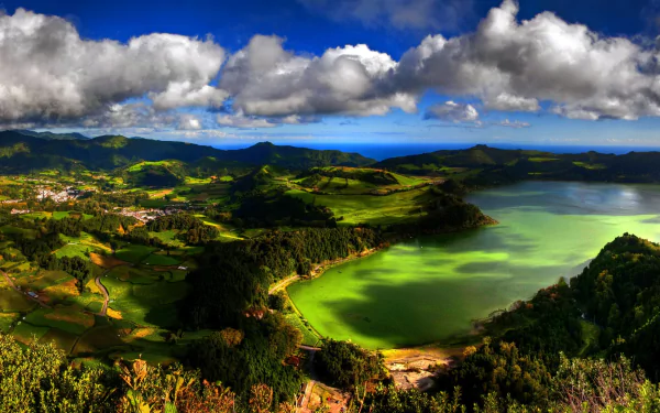 A panoramic view of a vibrant lake surrounded by lush forests and rolling hills under a dramatic sky with clouds, showcasing the stunning landscape of the Azores, Portugal.