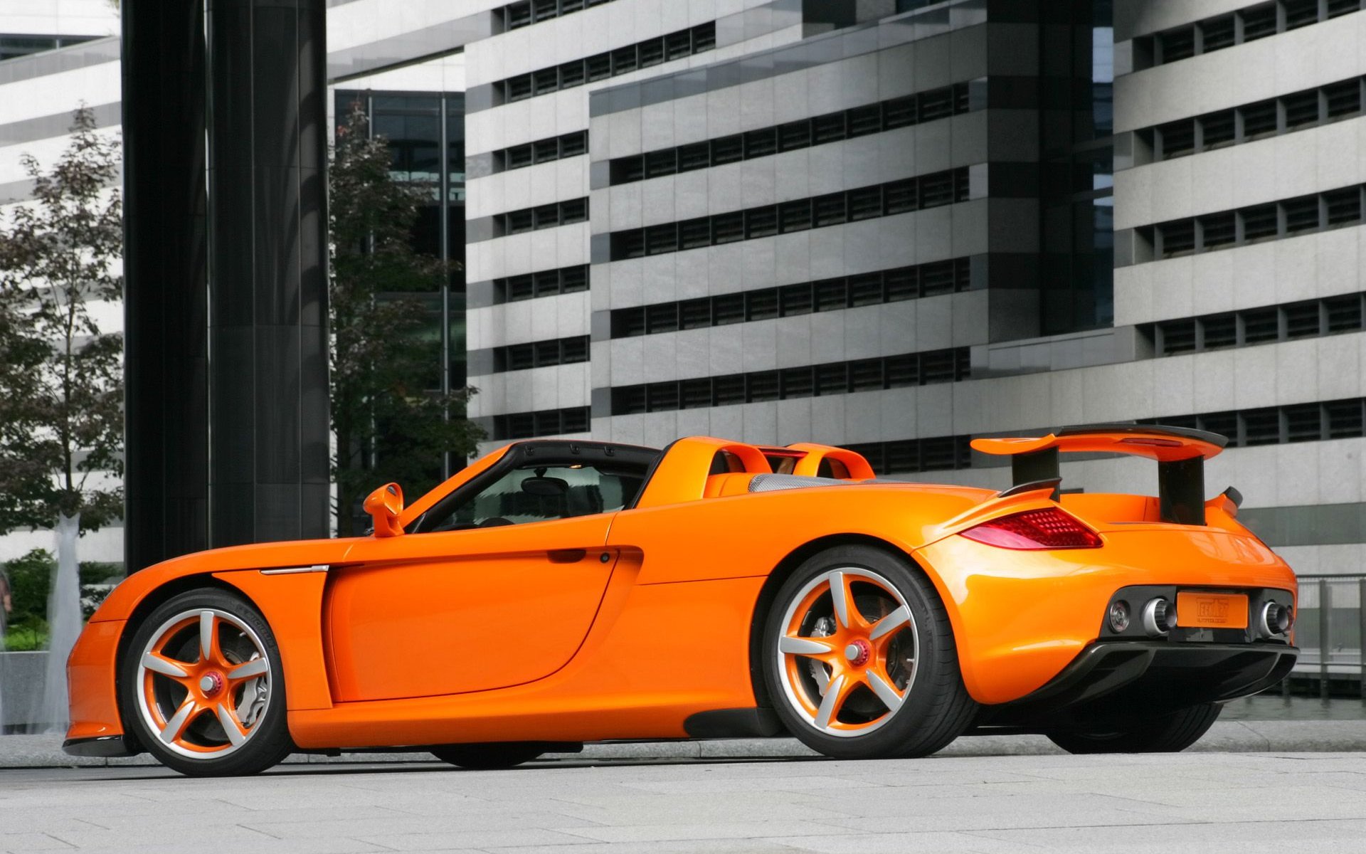 A striking orange Porsche Carrera GT parked against a modern architectural backdrop, showcasing its sleek design and dynamic presence in this HD desktop wallpaper.