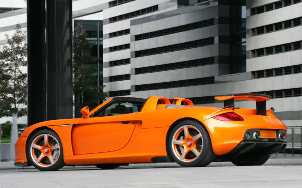 A striking orange Porsche Carrera GT parked against a modern architectural backdrop, showcasing its sleek design and dynamic presence in this HD desktop wallpaper.