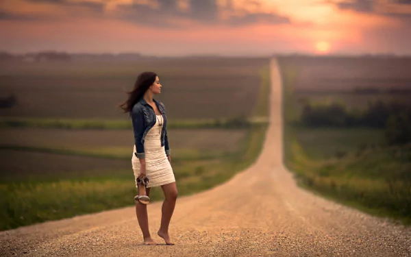 HD wallpaper of a beautiful woman standing barefoot on a long, straight road at sunset, holding her shoes in one hand, with a vast landscape stretching out to the horizon.