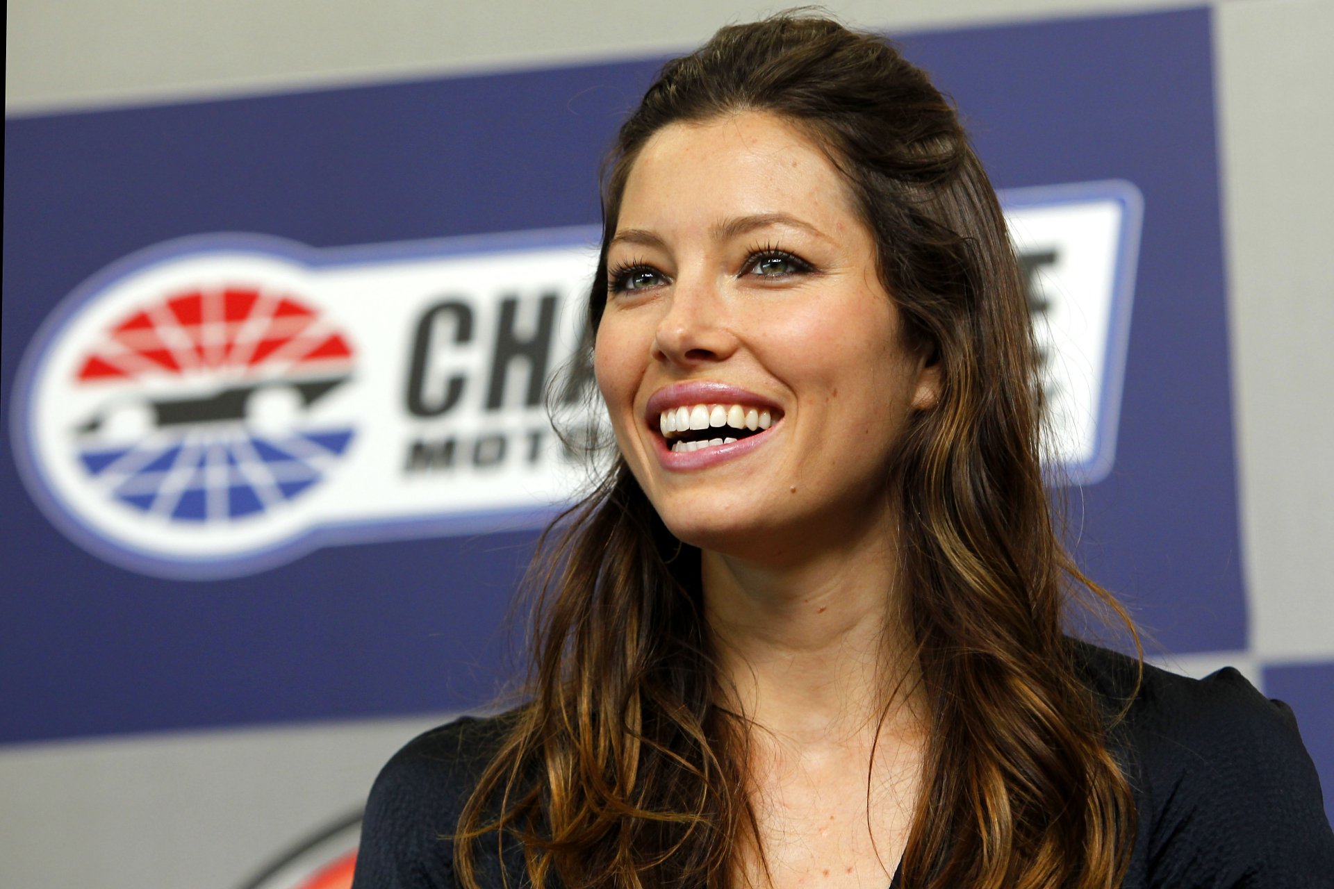 Smiling female celebrity with long brown hair against a motorsport logo backdrop — 4K Ultra HD PC desktop wallpaper/background.