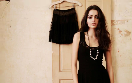 Brunette model Darla Baker in a black dress with necklace, standing beside a hanging skirt against a textured wall, HD desktop wallpaper background.