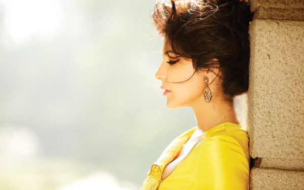 HD desktop wallpaper featuring celebrity Anushka Sharma in a yellow outfit, leaning against a stone wall with a serene outdoor background.