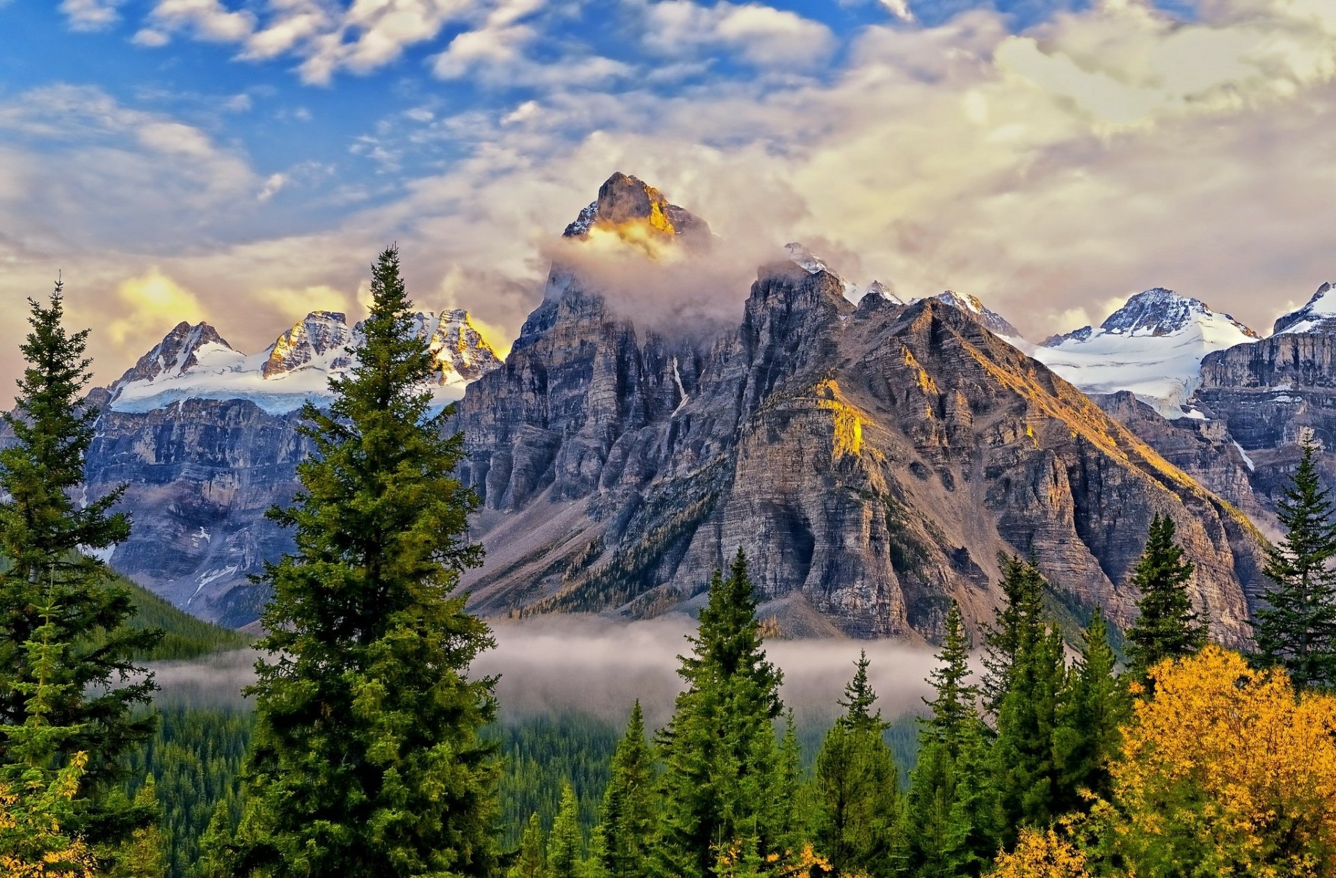HD PC desktop wallpaper featuring towering mountain peaks partly shrouded in fog and clouds, surrounded by dense evergreen trees under a vibrant blue sky.
