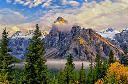 HD PC desktop wallpaper featuring towering mountain peaks partly shrouded in fog and clouds, surrounded by dense evergreen trees under a vibrant blue sky.