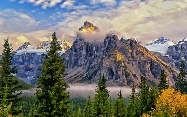 HD PC desktop wallpaper featuring towering mountain peaks partly shrouded in fog and clouds, surrounded by dense evergreen trees under a vibrant blue sky.