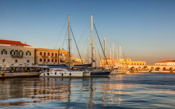 HD desktop wallpaper of Ermoupoli harbor at sunset, featuring sailboats docked alongside historic man-made buildings reflecting on calm waters.