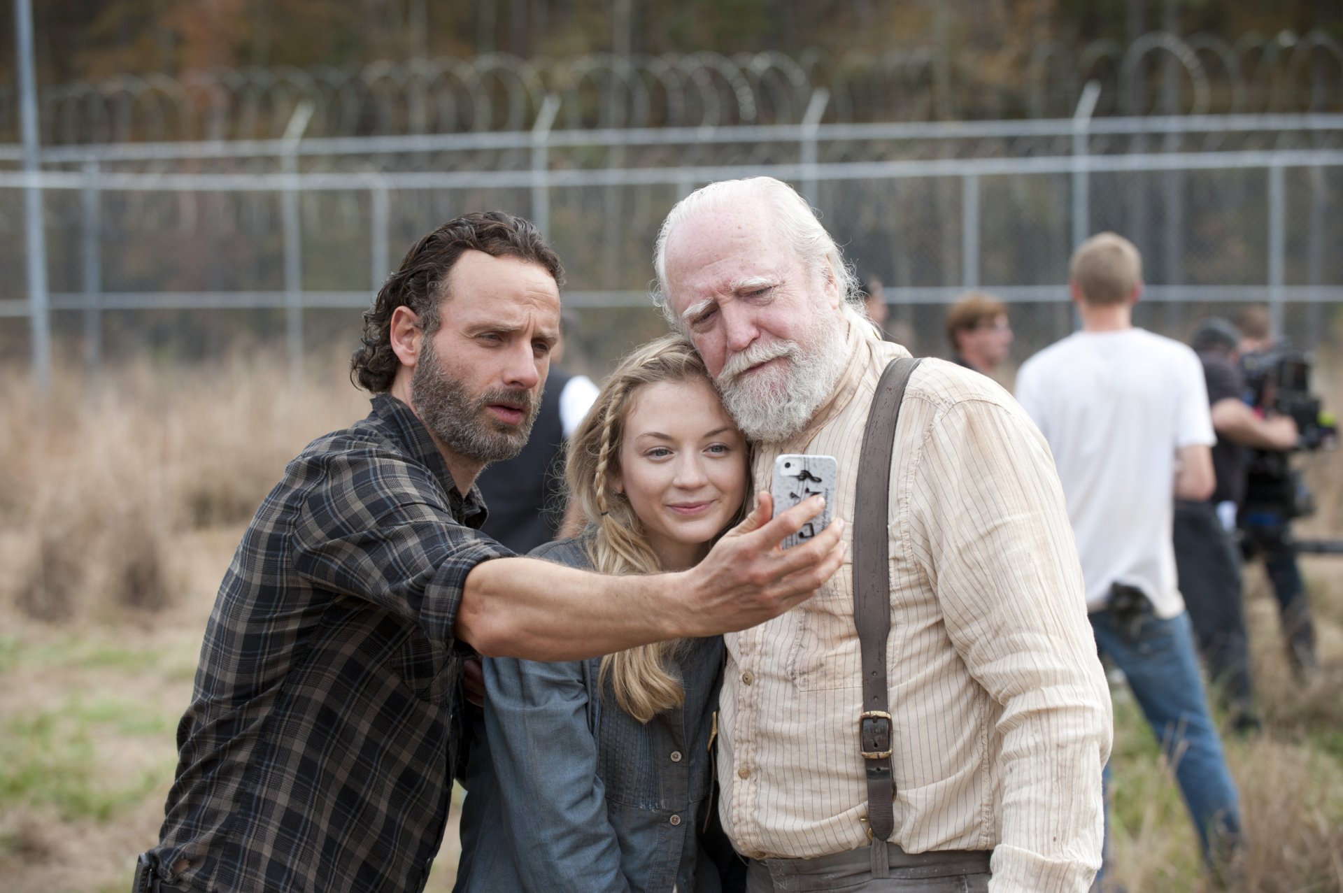 A scene from the TV show The Walking Dead, featuring characters Rick, a young woman, and an older man, taking a selfie against a backdrop of a fenced area.