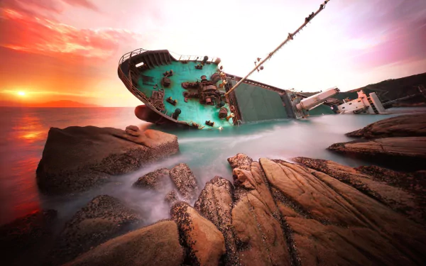 HD PC desktop wallpaper and background of a wrecked ship beached on jagged rocks at sunset, waves washing around the tilted green hull.