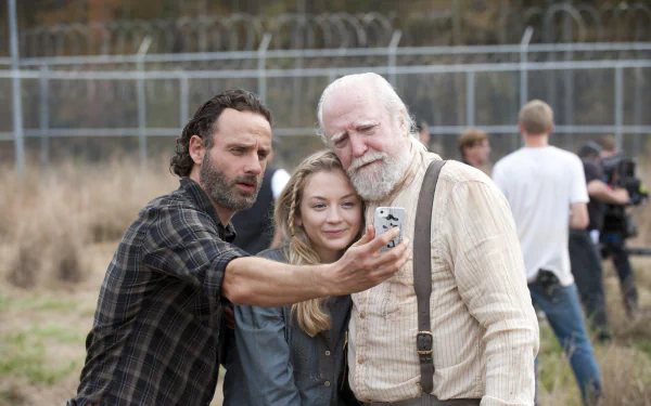 A scene from the TV show The Walking Dead, featuring characters Rick, a young woman, and an older man, taking a selfie against a backdrop of a fenced area.