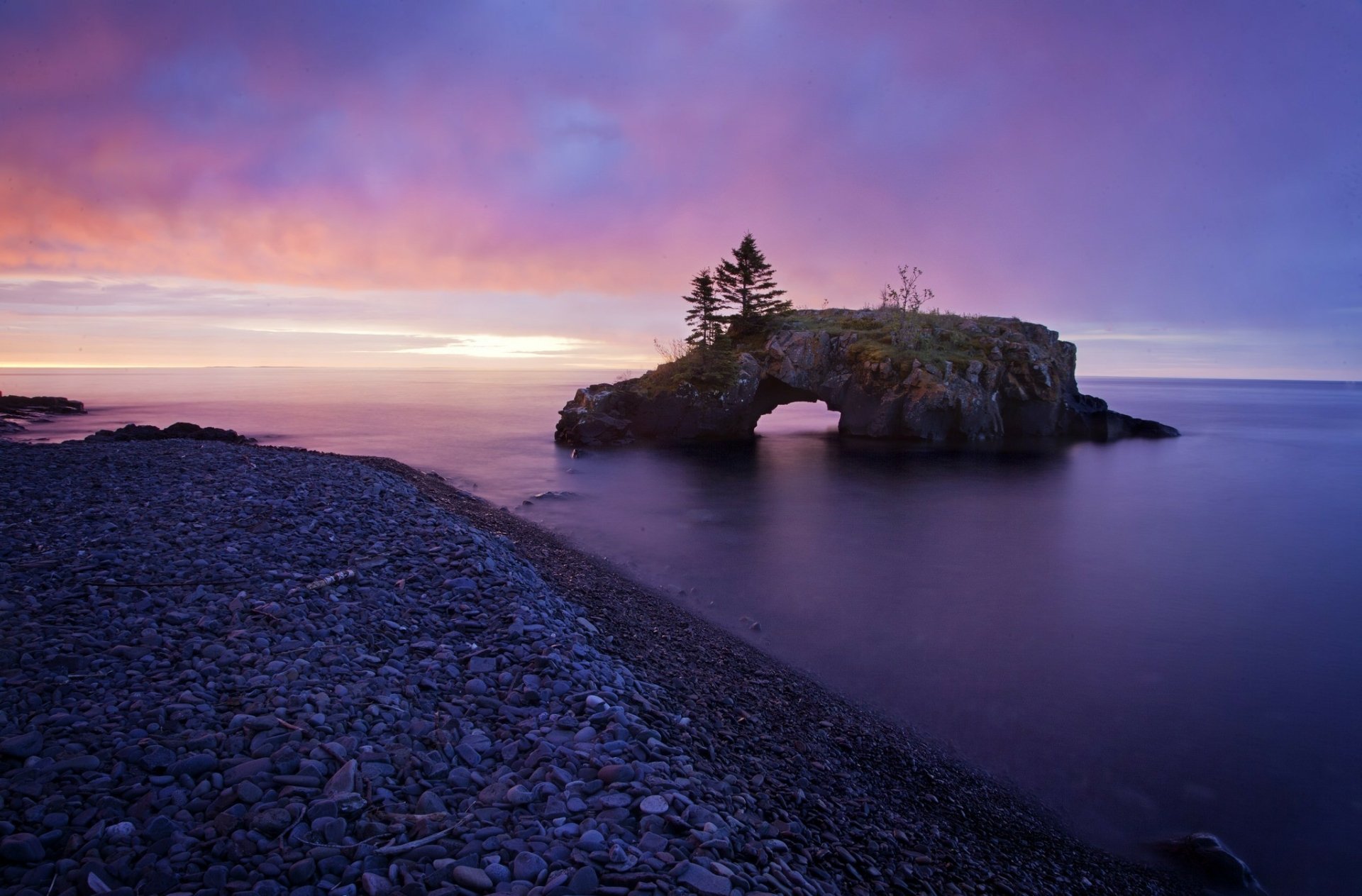 HD PC desktop wallpaper — serene seashore at twilight: pebble beach foreground, a small rocky arch island topped with trees, purple-pink sky reflecting on calm sea.