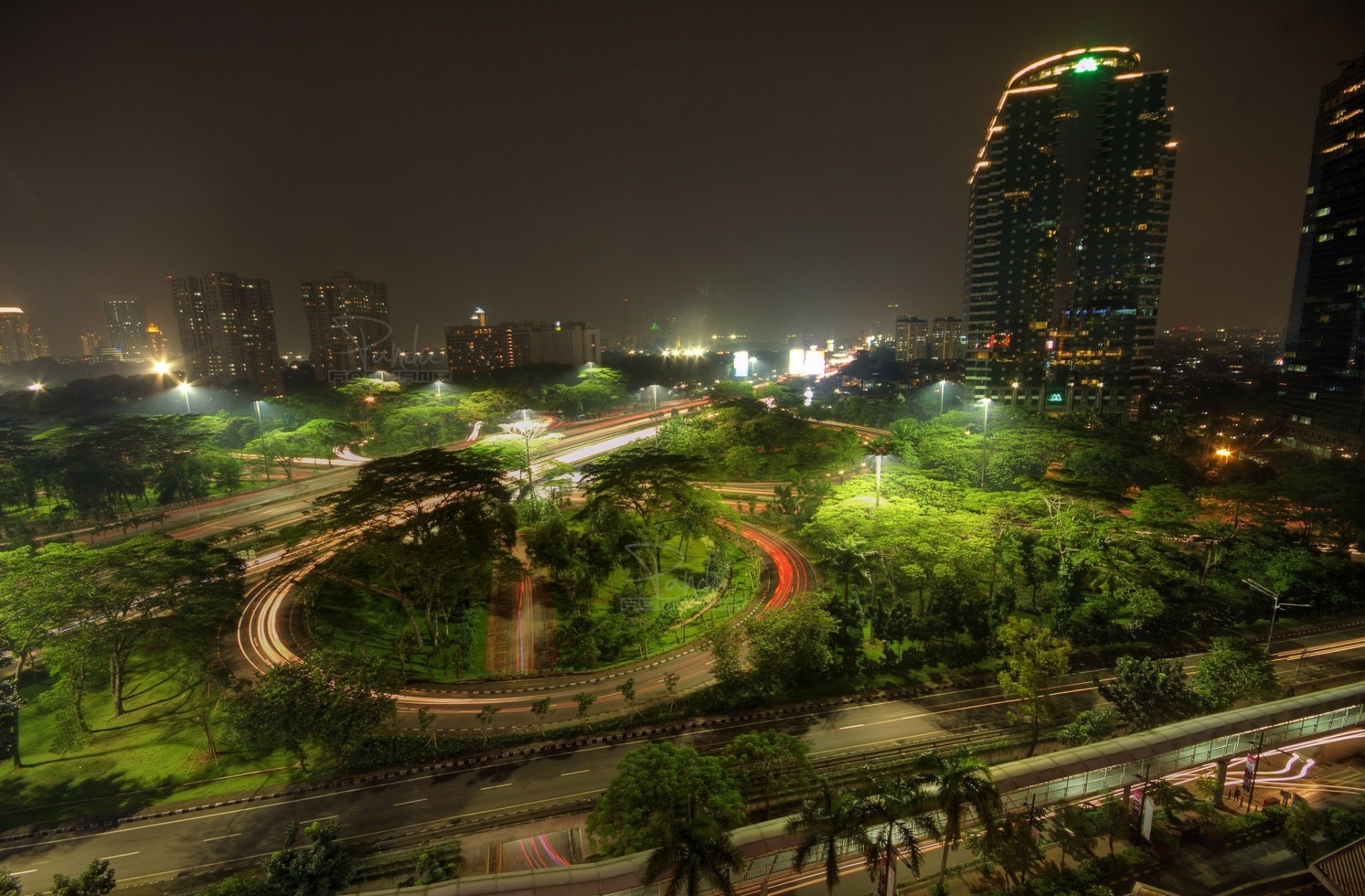 Nighttime view of Jakarta, Java, Indonesia, showcasing illuminated city streets, greenery, and tall buildings under a dark sky.