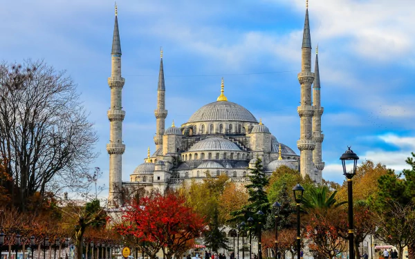 A stunning view of the Sultan Ahmed Mosque in Istanbul, Turkey, showcasing its iconic architecture and surrounding vibrant trees against a beautifully cloudy sky.