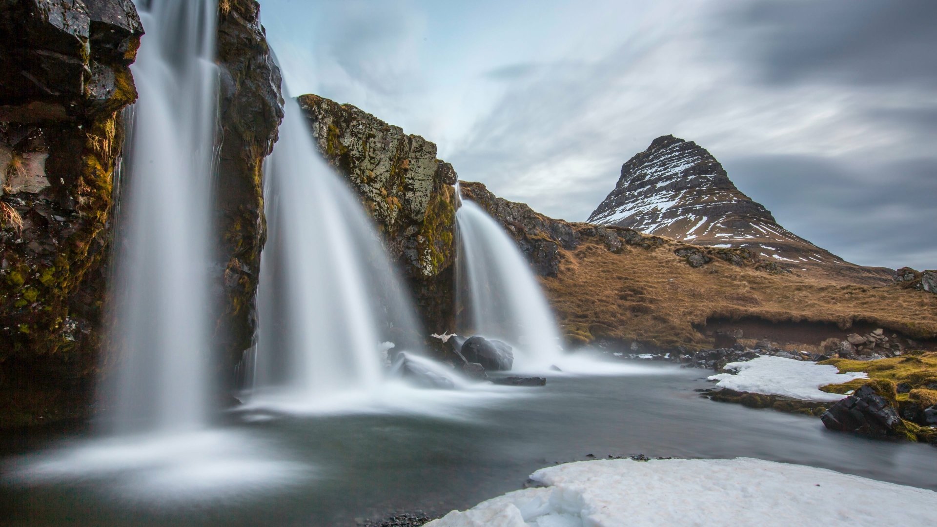 4K Ultra HD PC desktop wallpaper showing a nature waterfall scene: silky cascades over mossy cliffs, a river with ice patches and a distant snow-capped peak under cloudy skies.