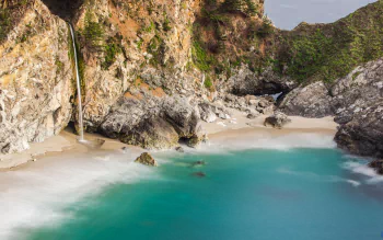 A serene seascape featuring a rocky shoreline, gentle waterfall, and calm turquoise waters, captured in stunning 4K Ultra HD for a vibrant PC desktop wallpaper.