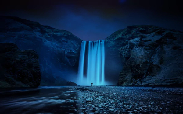 A solitary figure stands before the majestic Skógafoss Waterfall, surrounded by dark cliffs and a serene landscape, creating a stunning nature-inspired HD wallpaper.