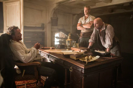 Captain Flint from Black Sails, played by Toby Stephens, engages with two crew members around a wooden table in a dimly lit pirate hideout, capturing the show's intense atmosphere.