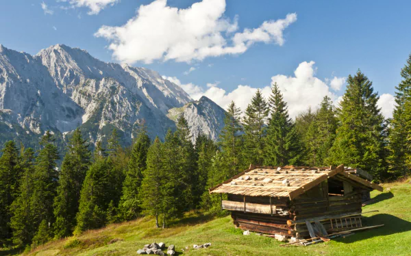 4K Ultra HD PC desktop wallpaper/background: a man-made wooden cabin on a sunlit alpine meadow, framed by pine trees and rugged mountains under a bright blue sky.