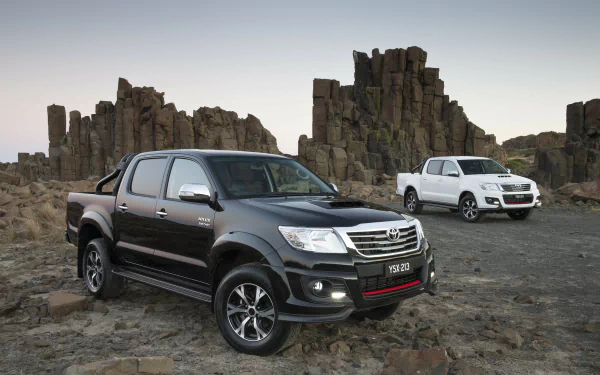 HD desktop wallpaper featuring two Toyota Hilux vehicles parked on rocky terrain with rugged rock formations in the background.