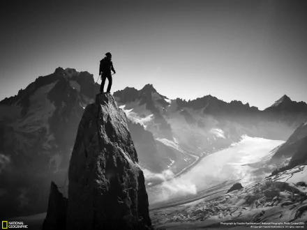 A climber stands atop a jagged peak, surrounded by majestic snow-covered mountains and a sweeping valley. This striking black-and-white image captures the essence of winter sports and adventure.