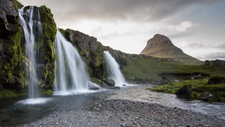 A stunning waterfall cascades over lush greenery, flowing into a serene river, with a majestic mountain rising in the background. This HD image captures the beauty of nature perfectly.