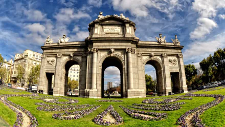 HD desktop wallpaper showcasing the man-made Puerta de Alcalá monument under a vibrant sky with decorative flowerbeds in the foreground.