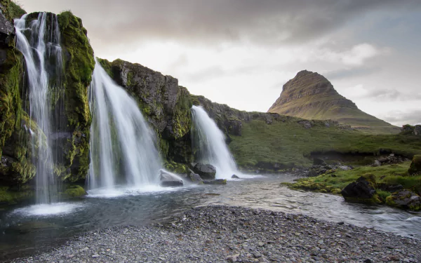 A stunning waterfall cascades over lush greenery, flowing into a serene river, with a majestic mountain rising in the background. This HD image captures the beauty of nature perfectly.