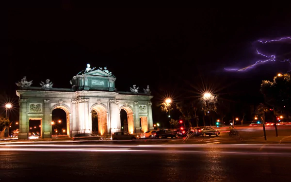 Night view of the illuminated Puerta de Alcalá with lightning in the sky, captured as an HD man-made desktop wallpaper and background.