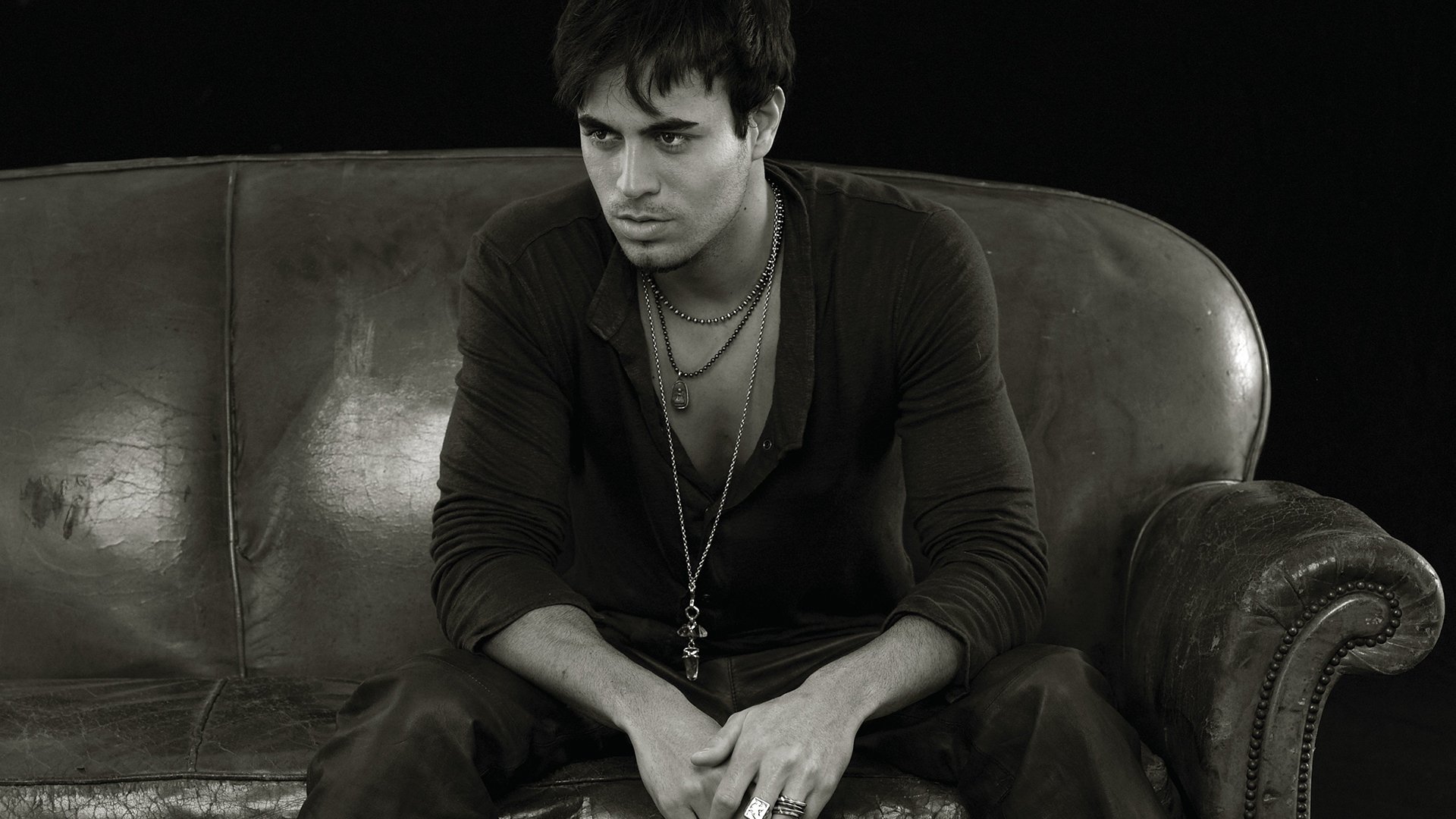 Black and white HD wallpaper of Enrique Iglesias sitting on a leather couch, exuding a contemplative expression. Tags: music, Enrique Iglesias.