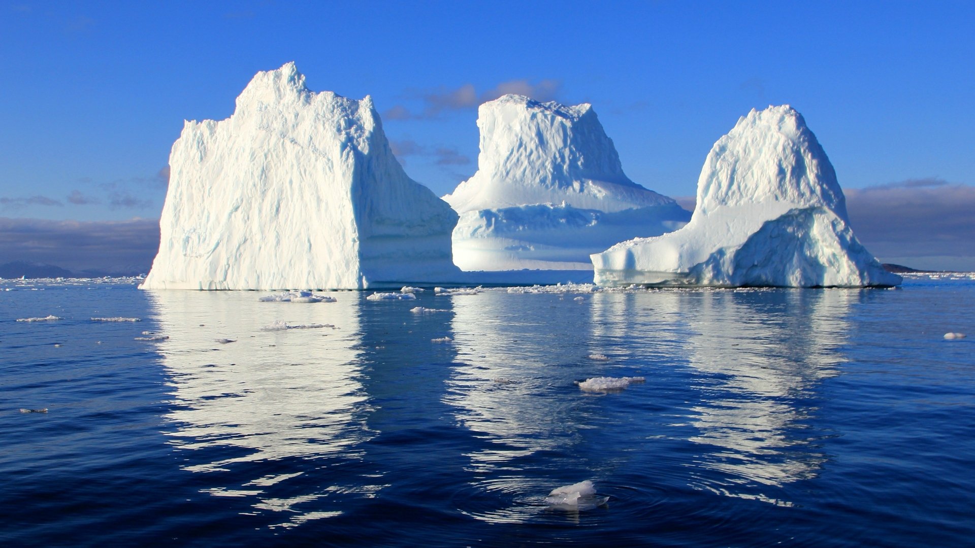 4K Ultra HD PC desktop wallpaper and background: serene nature scene of three majestic icebergs floating on a calm blue sea, their crisp reflections mirrored beneath a clear sky.