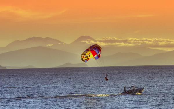 4K Ultra HD desktop wallpaper showing a vibrant parasailing scene over calm ocean waters with mountains and a colorful sunset sky in the background.