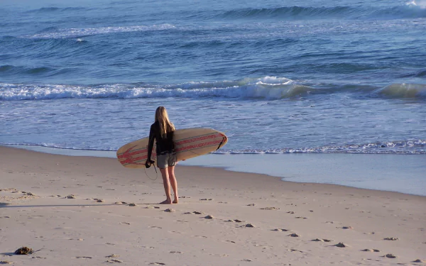 Surfer holding a surfboard stands on a sandy beach facing ocean waves, captured in HD for a vibrant desktop wallpaper showcasing beach and surfing sports.