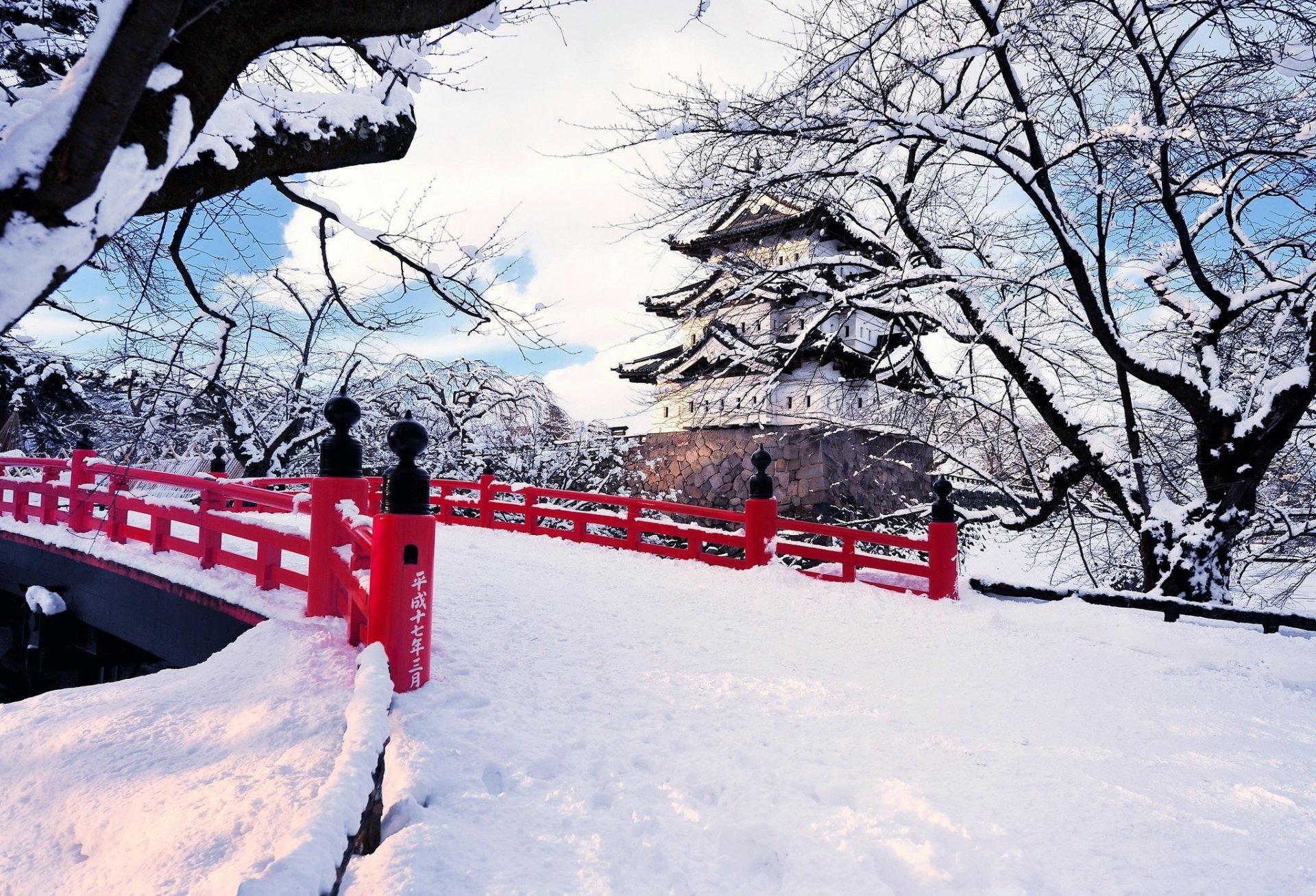 Snow-covered Hirosaki Castle and red bridge in Aomori Prefecture, framed by bare trees under a bright winter sky.