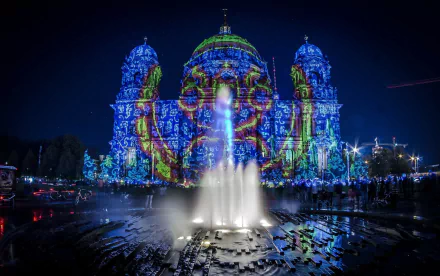 A vibrant nighttime scene in Berlin featuring a beautifully illuminated fountain, with colorful lights projected onto nearby architectural structures. An enchanting urban backdrop for any desktop.