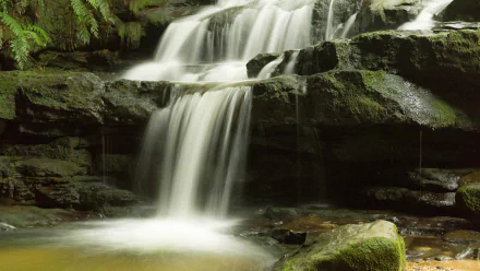 A 4K Ultra HD desktop wallpaper featuring a serene waterfall cascading over moss-covered rocks in a lush, green natural setting.
