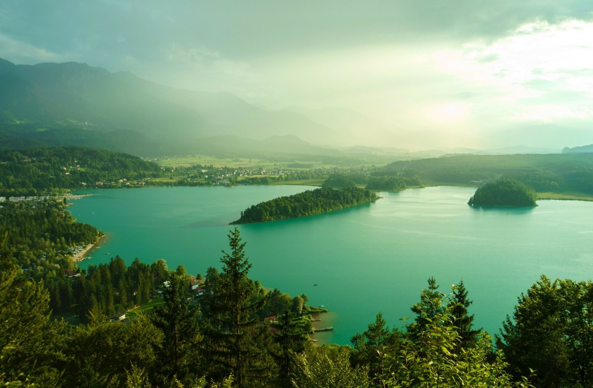 A serene HD desktop wallpaper featuring Austria’s lake surrounded by lush green forests and misty mountains under a cloudy sky.