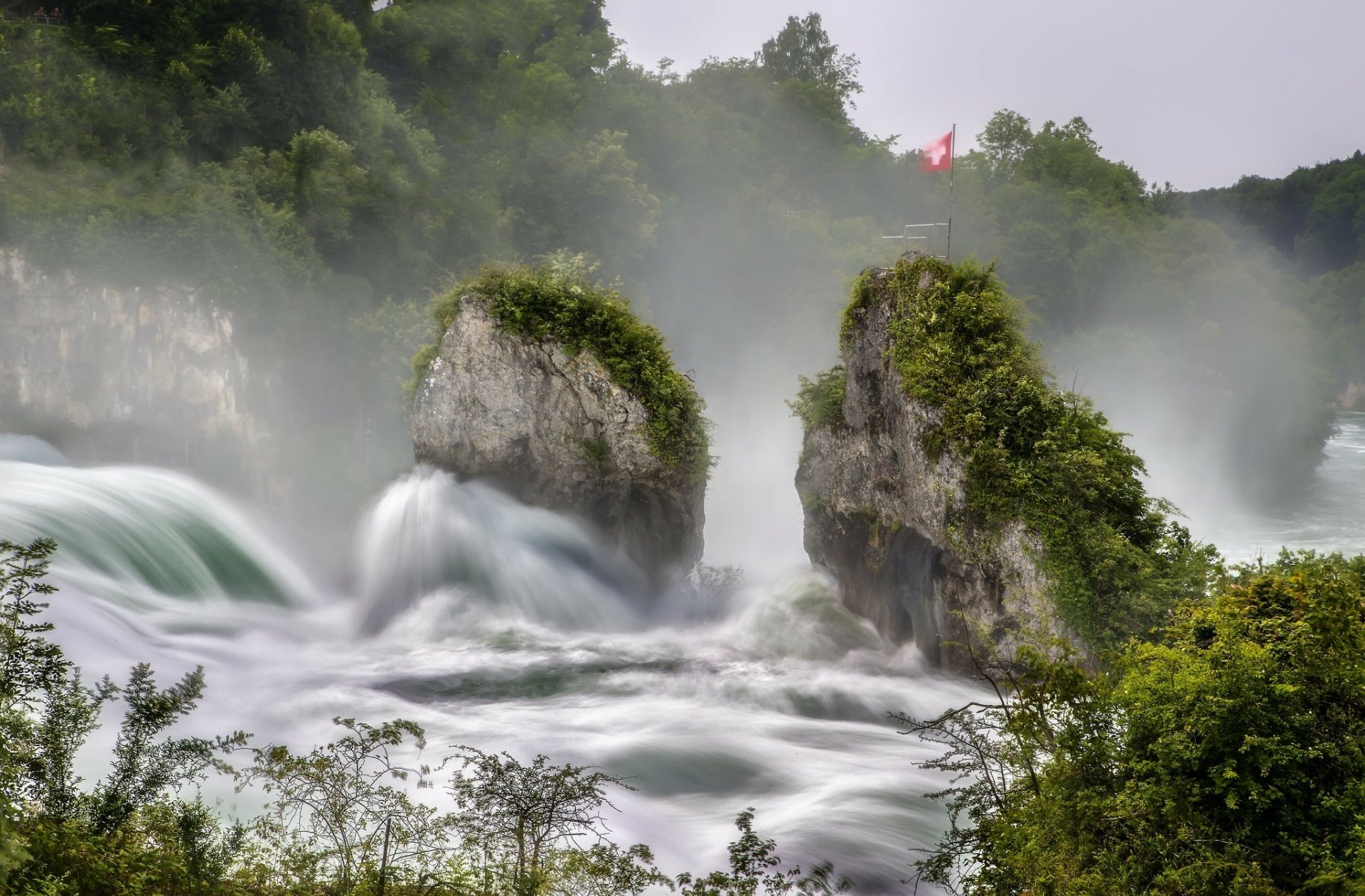 Powerful torrent of water cascades around moss-covered rocks with the Swiss flag above, set in lush greenery, capturing the raw energy and beauty of nature in HD.