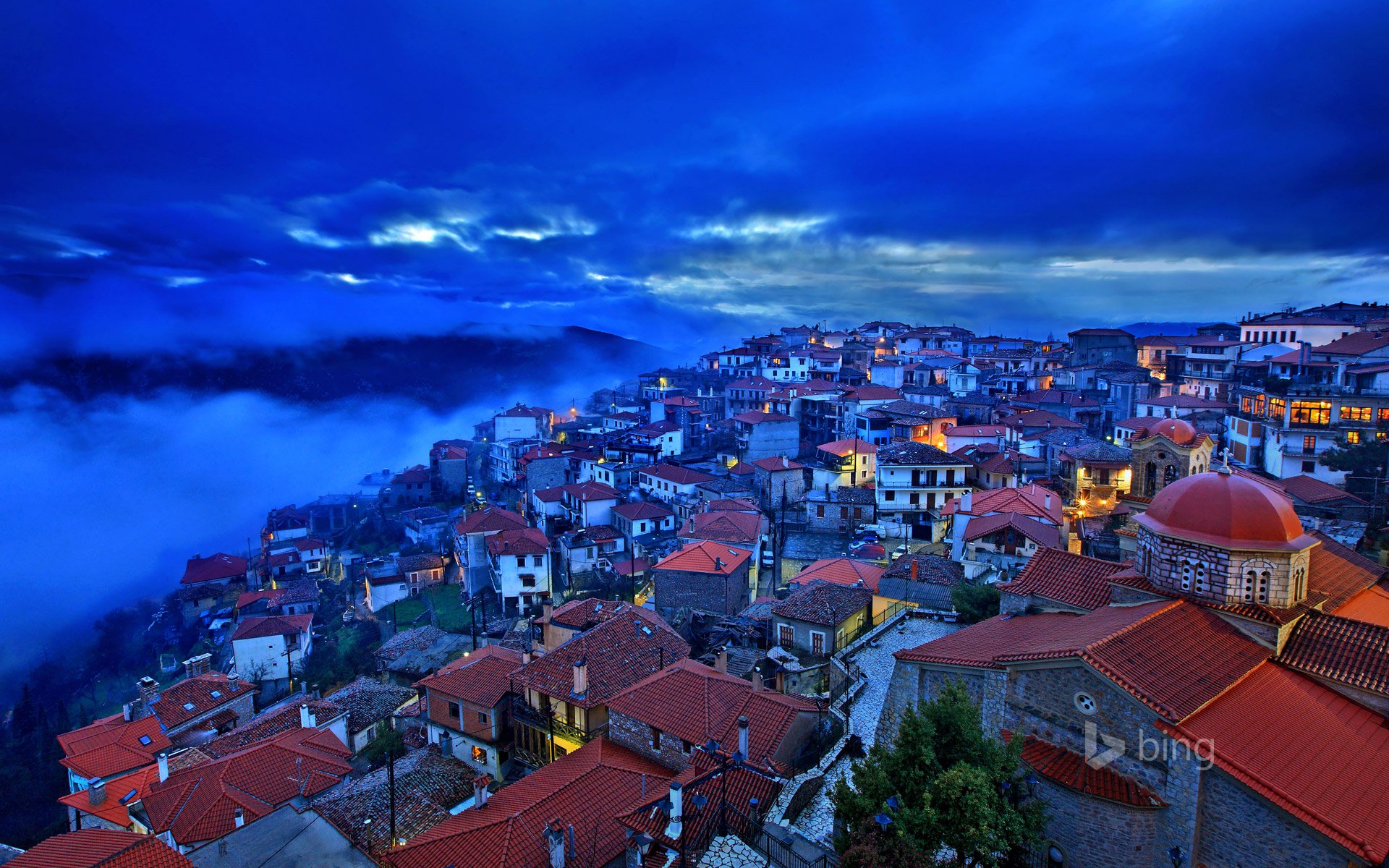 A breathtaking HD desktop wallpaper featuring the man-made town of Arachova, Greece, nestled on a hillside under a dramatic blue sky with glowing lights.