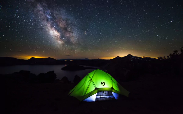 A vibrant green tent glows under the stunning Milky Way, set against a serene lakeside backdrop, showcasing the beauty of camping and astrophotography in high definition.