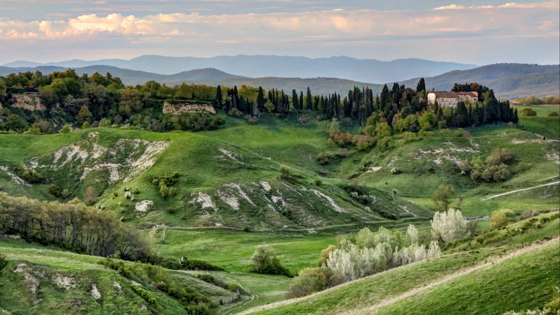 Nature landscape 2K Quad HD PC desktop wallpaper: rolling green hills, groves of cypress and poplar, winding paths and layered blue mountains beneath a soft, cloud-speckled sky.