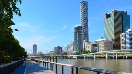 A vibrant Brisbane skyline featuring the Infinity Tower, Kurilpa Bridge, and a scenic walkway along the river, showcasing modern skyscrapers and city life against a clear blue sky.