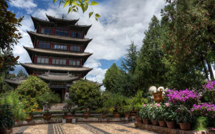 A vibrant 4K image of the Lijiang Pagoda temple in Yunnan, China, surrounded by lush greenery and colorful flowers under a partly cloudy sky.