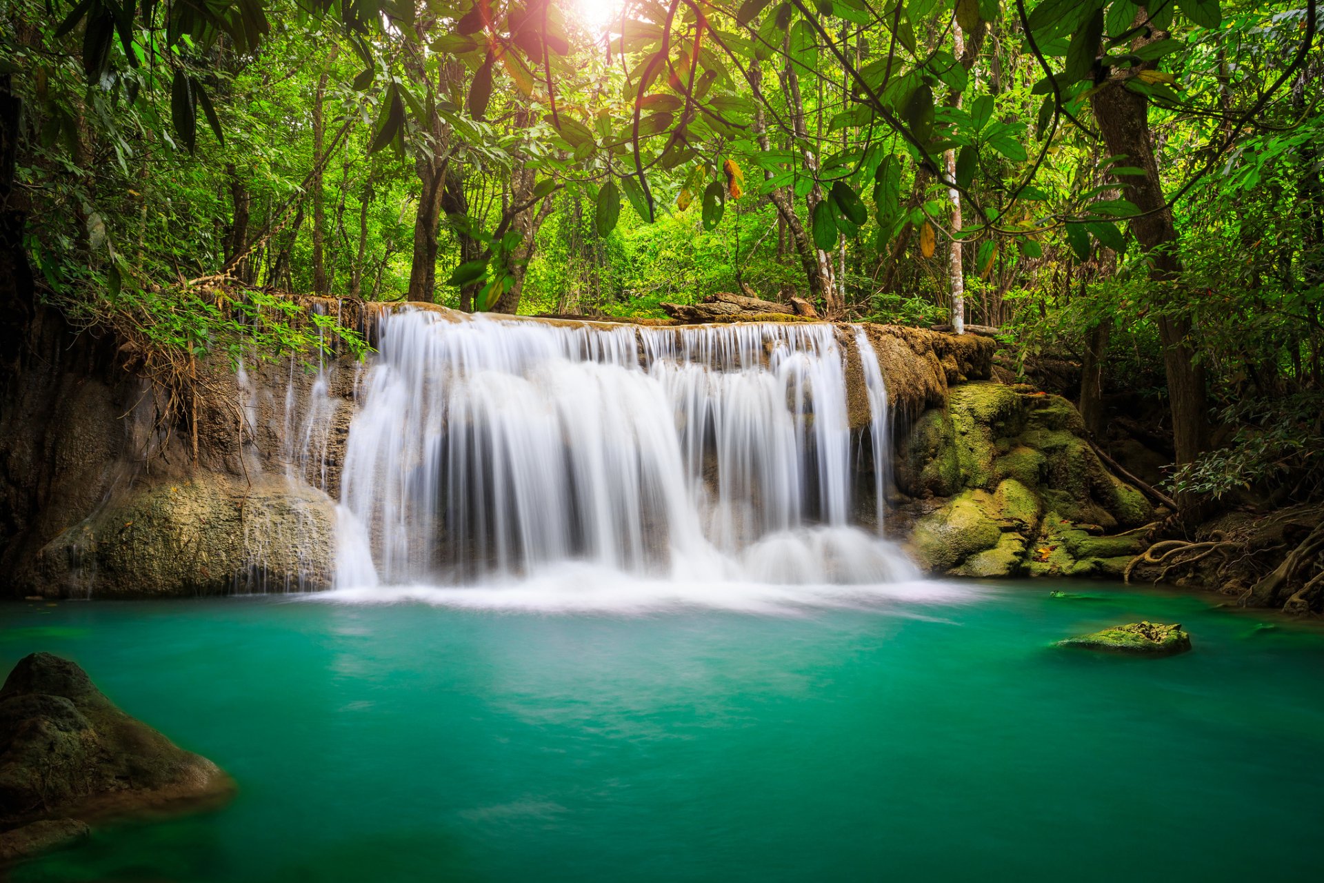 Lush green forest surrounds a serene waterfall cascading into turquoise water in Thailand, captured in vibrant HD for a stunning desktop wallpaper.