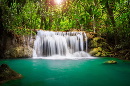 Lush green forest surrounds a serene waterfall cascading into turquoise water in Thailand, captured in vibrant HD for a stunning desktop wallpaper.