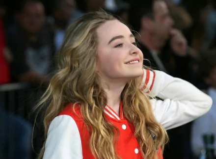A high-definition desktop wallpaper featuring popular celebrity Sabrina Carpenter, smiling and wearing a red and white varsity jacket, with a lively crowd in the background.