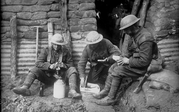 Three soldiers in World War I, seated in a trench, engage with their equipment. The image captures a moment of camaraderie amid the realities of military life. HD wallpaper background.