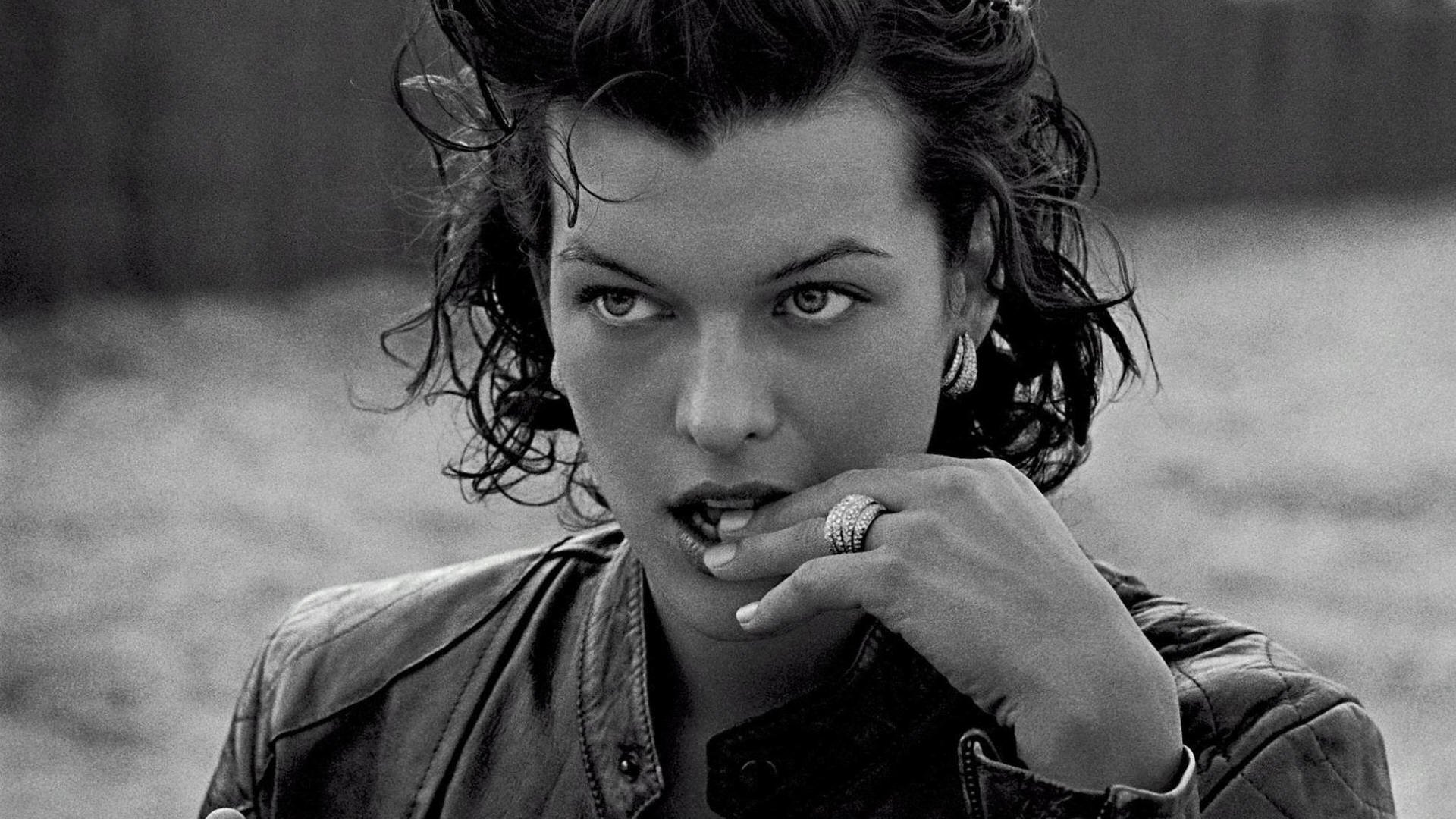 Black and white HD desktop wallpaper featuring celebrity Milla Jovovich with wet hair, wearing a leather jacket, gazing thoughtfully while biting her finger.