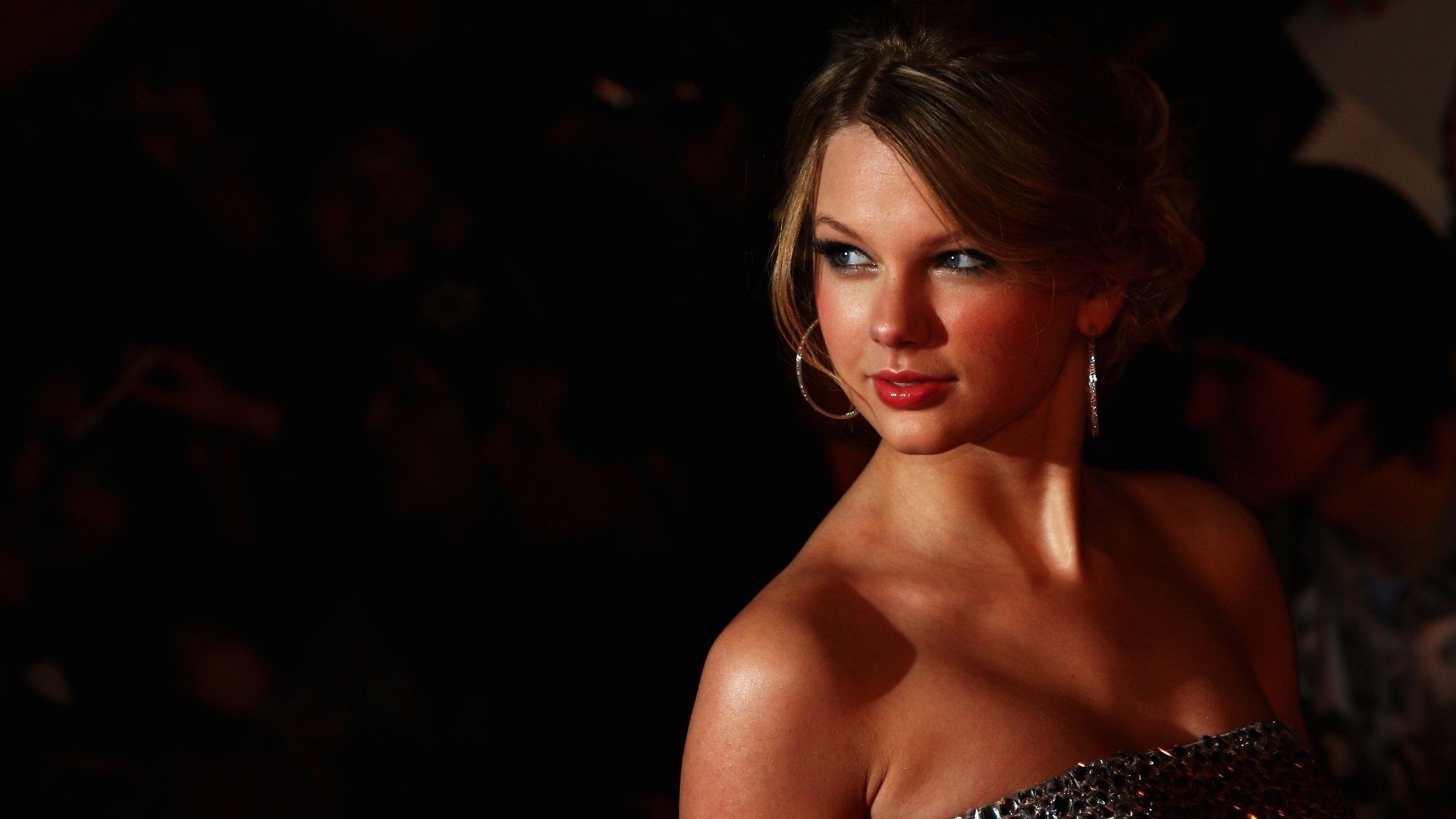 A glamorous Taylor Swift, an American singer with blonde hair, poses elegantly in a shimmering outfit against a dark backdrop, highlighted in this stunning 4K Ultra HD wallpaper.