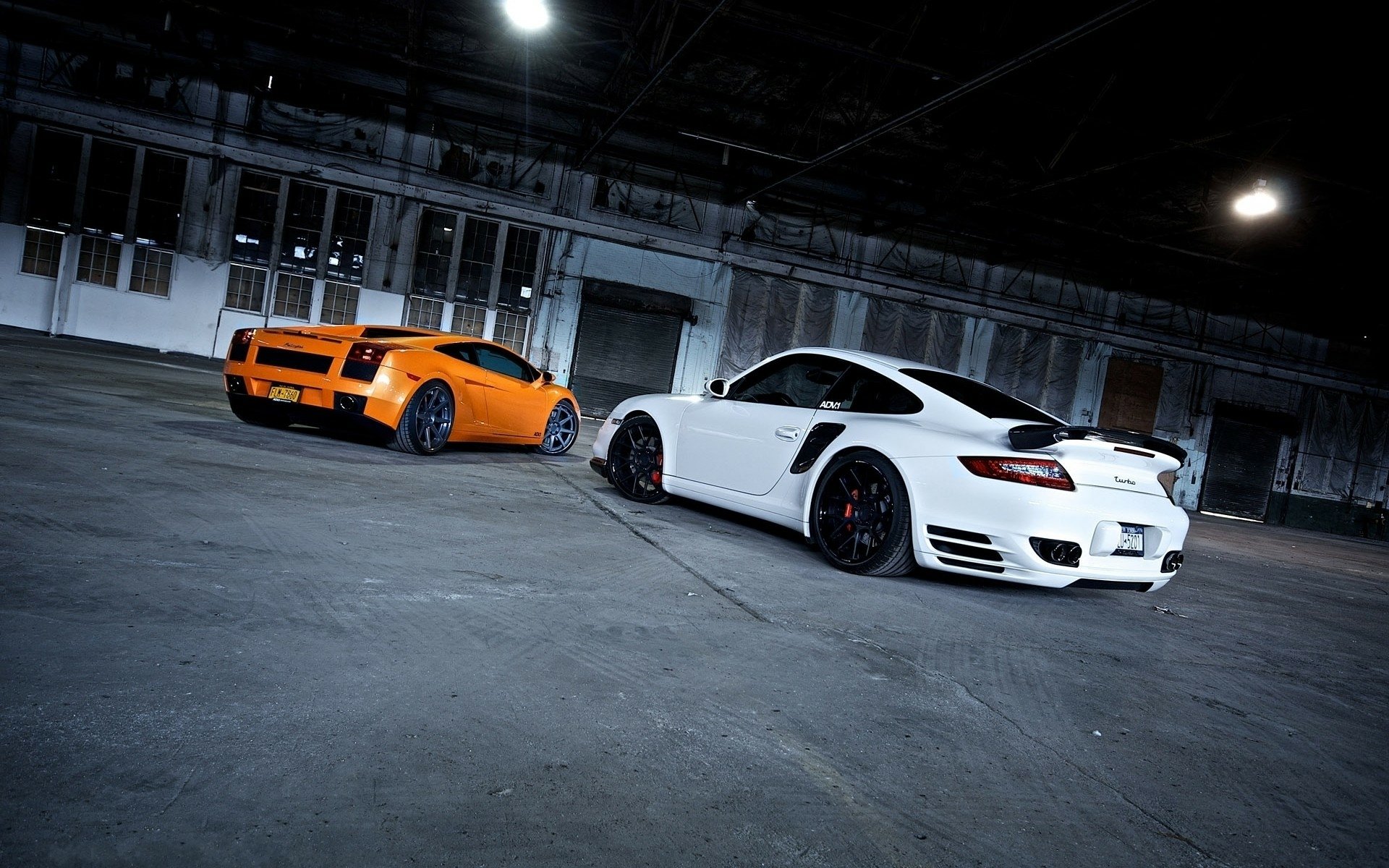 HD PC desktop wallpaper featuring a white Porsche and an orange Lamborghini sports car parked in an industrial-style garage.