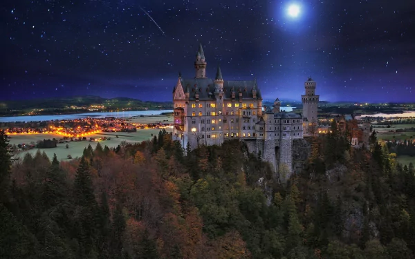 A stunning night scene of Neuschwanstein Castle in Germany, illuminated under a starry sky, with a bright moon and a meteor streaking across the horizon. A captivating desktop wallpaper.